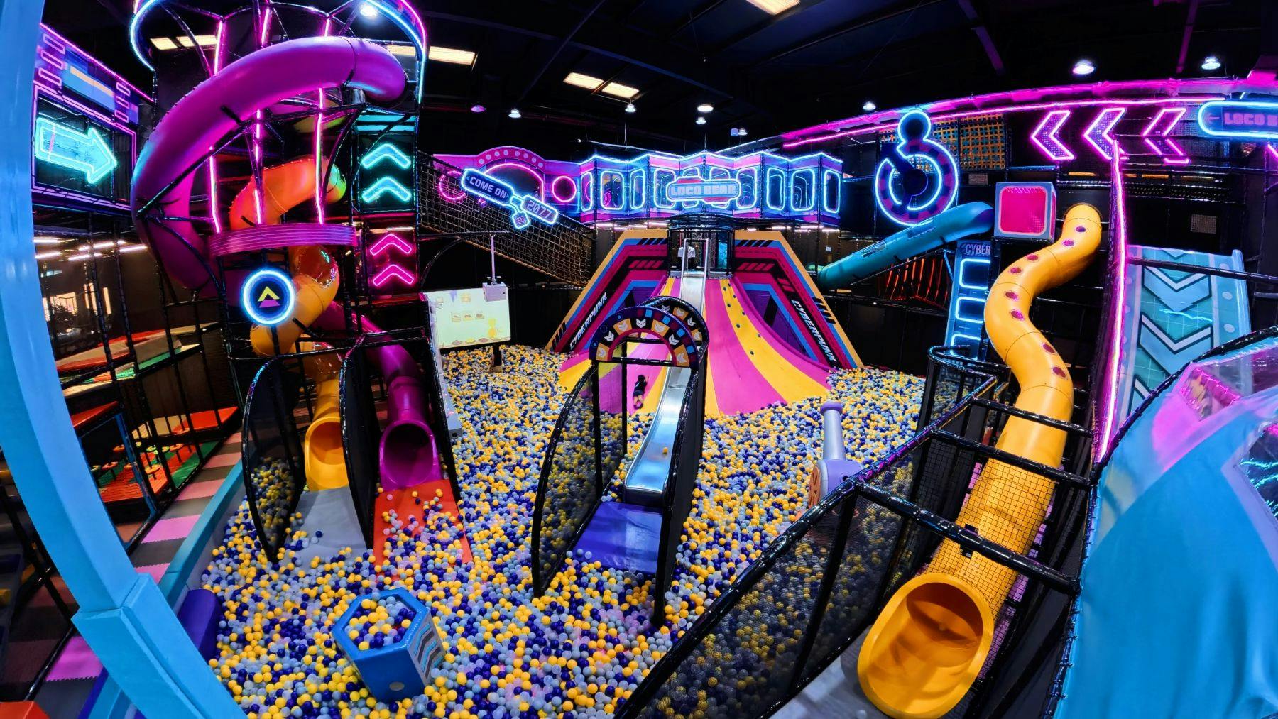 Ένας πολύχρωμος εσωτερικός χώρος παιχνιδιού με τσουλήθρες, σφαιρικούς λάκκους και φώτα νέον σε ένα ζωντανό, φουτουριστικό θέμα.