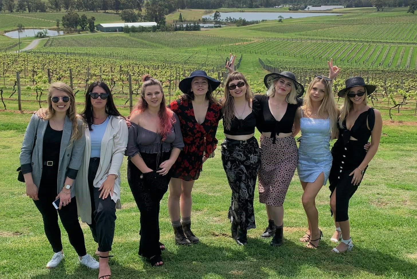 A group of six women posing together on a grassy field with vineyards and a landscape in the background.
