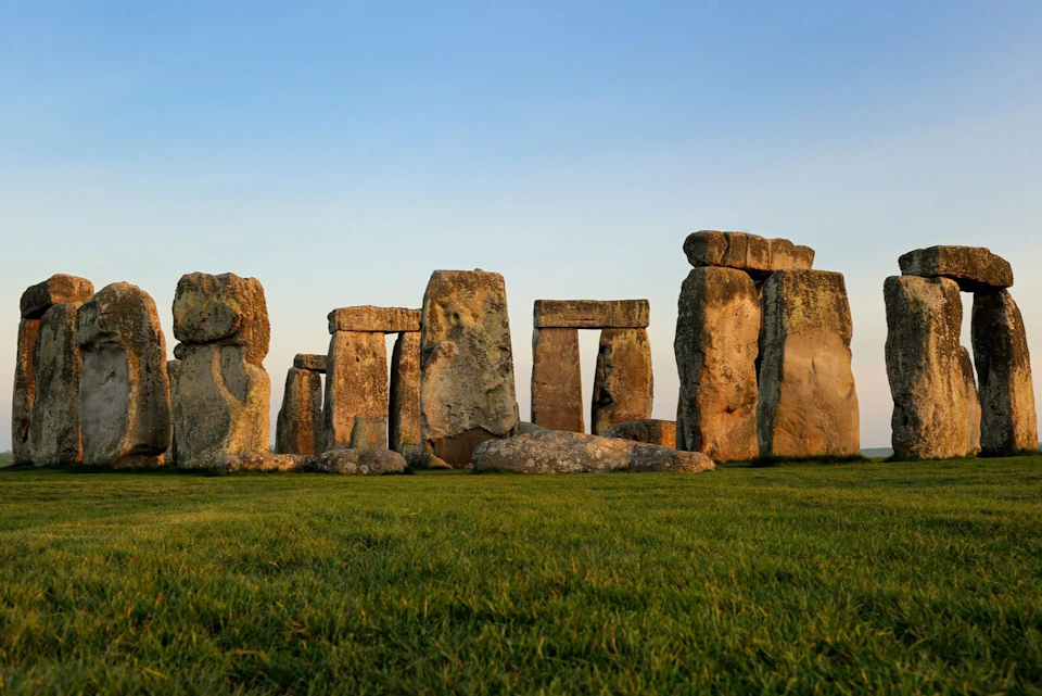Stonehenge, an ancient stone circle monument, stands on a grassy field under a clear blue sky at sunset.
