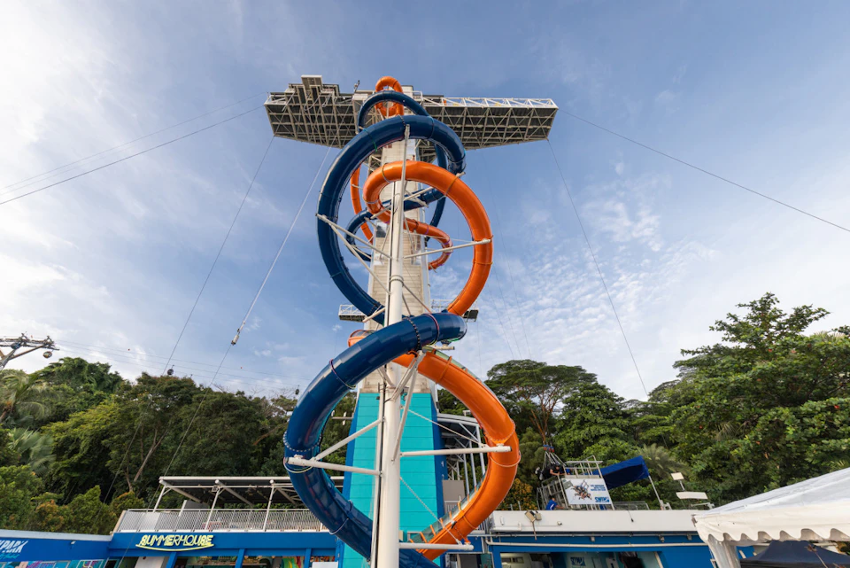 A tall water slide structure with intertwined blue and orange slides under a clear sky, surrounded by trees.