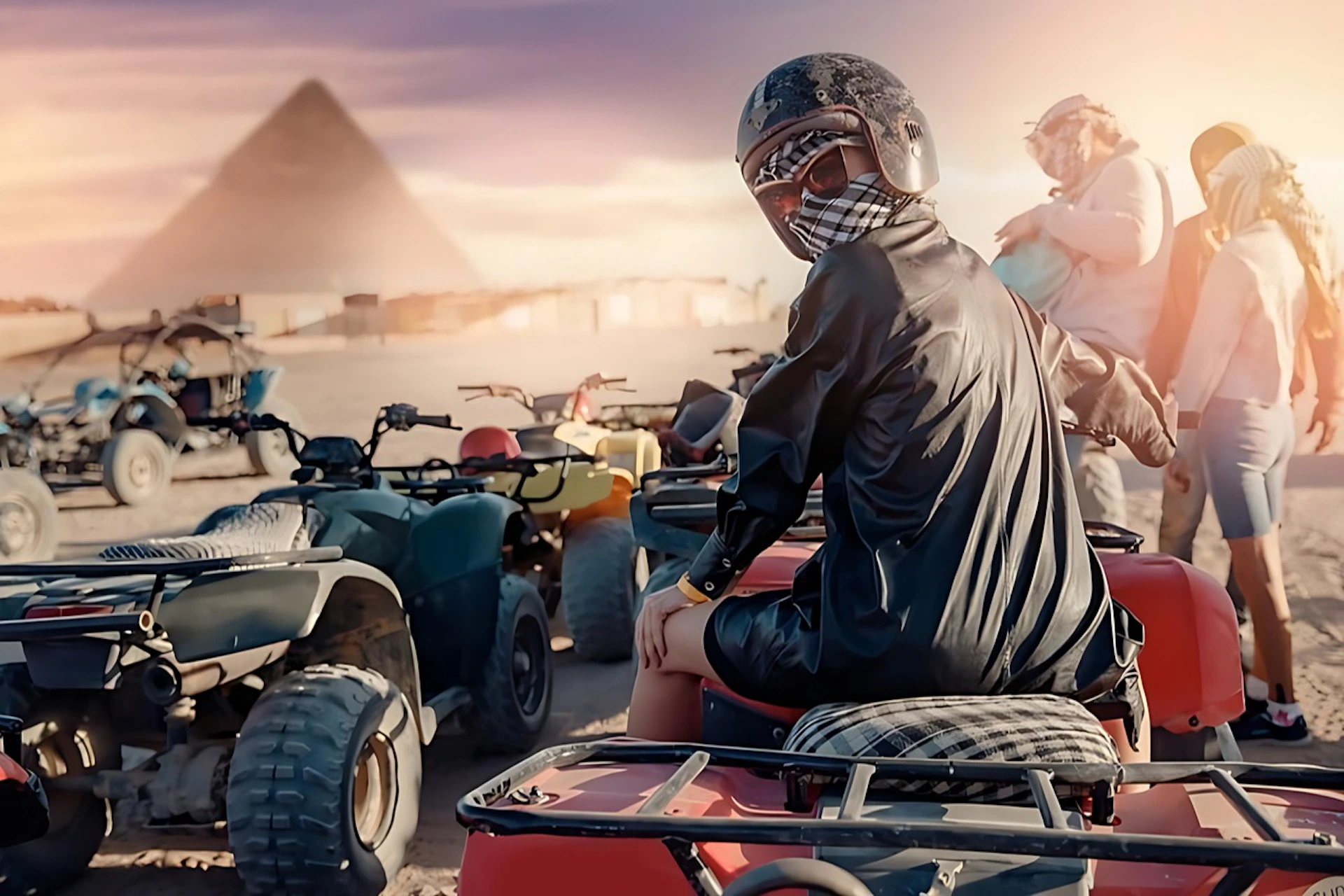 Person in protective gear sitting on an ATV, with several other ATVs around and a pyramid visible in the background.