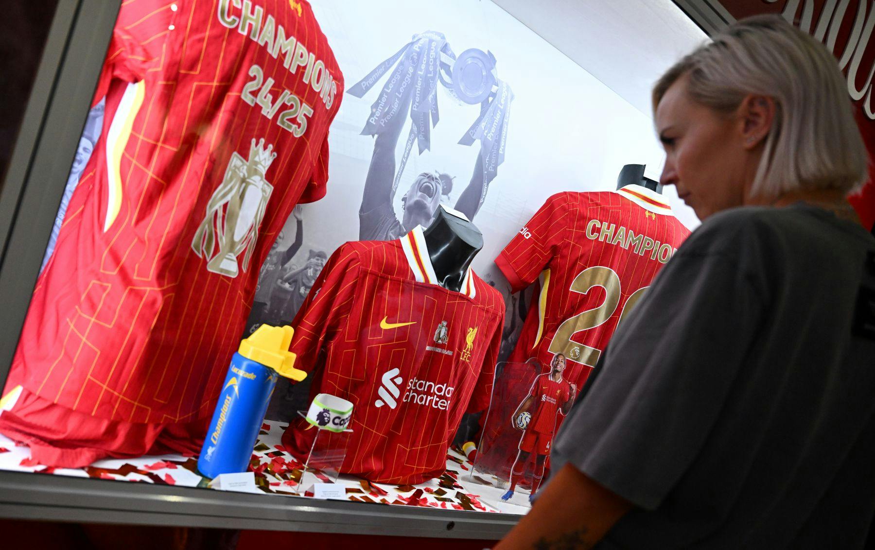 한 사람이 트로피를 수상한 사진을 배경으로 빨간색 축구 유니폼이 있는 디스플레이 케이스를 바라보고 있습니다.