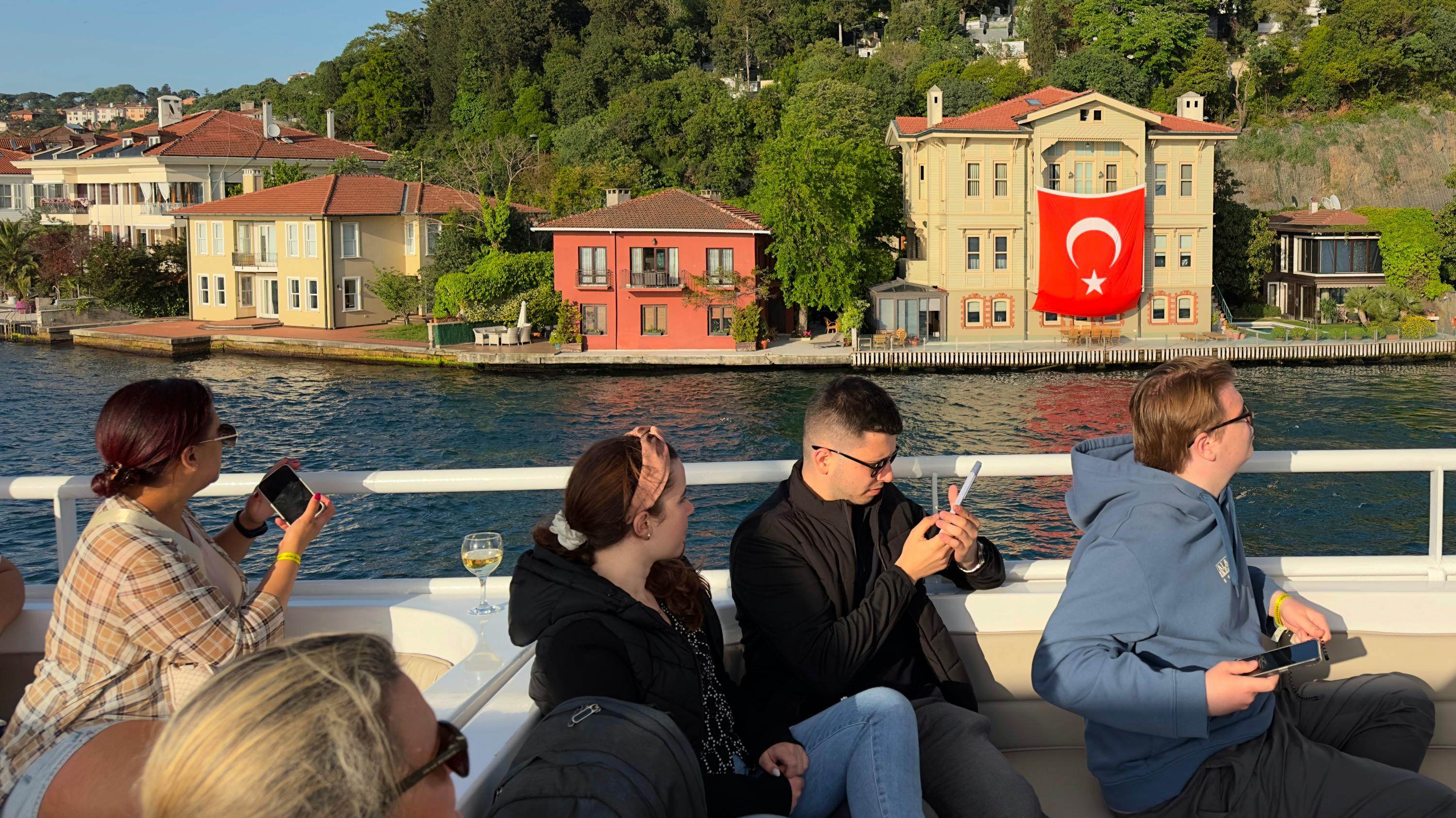 People sitting on a boat in front of waterfront houses, one with a Turkish flag, on a sunny day.