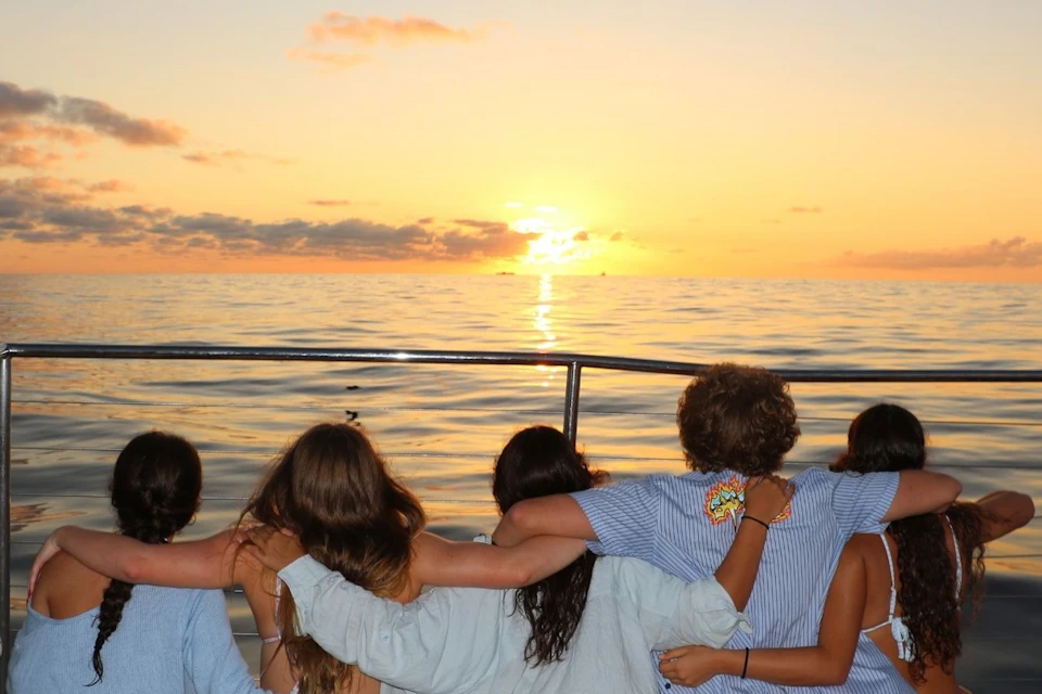 A group of people sitting together on a boat, arms around each other, watching a sunset over calm ocean waters.