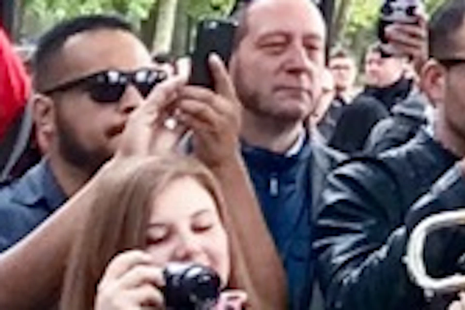 Musicians in red uniforms and bearskin hats play brass instruments; a crowd of onlookers takes photos behind a barrier.