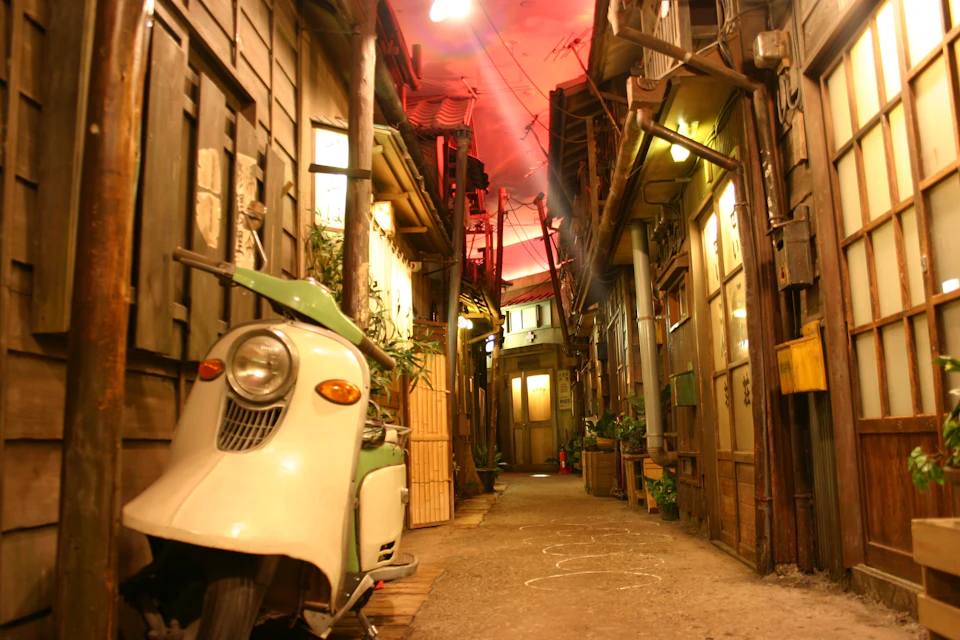 A narrow alleyway with old wooden buildings, a vintage scooter on the left, and a red-illuminated sky above.