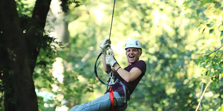 Pessoa a fazer tirolesa numa floresta, usando um capacete e um arnês, sorrindo com uma expressão de entusiasmo. Árvores e vegetação em fundo.