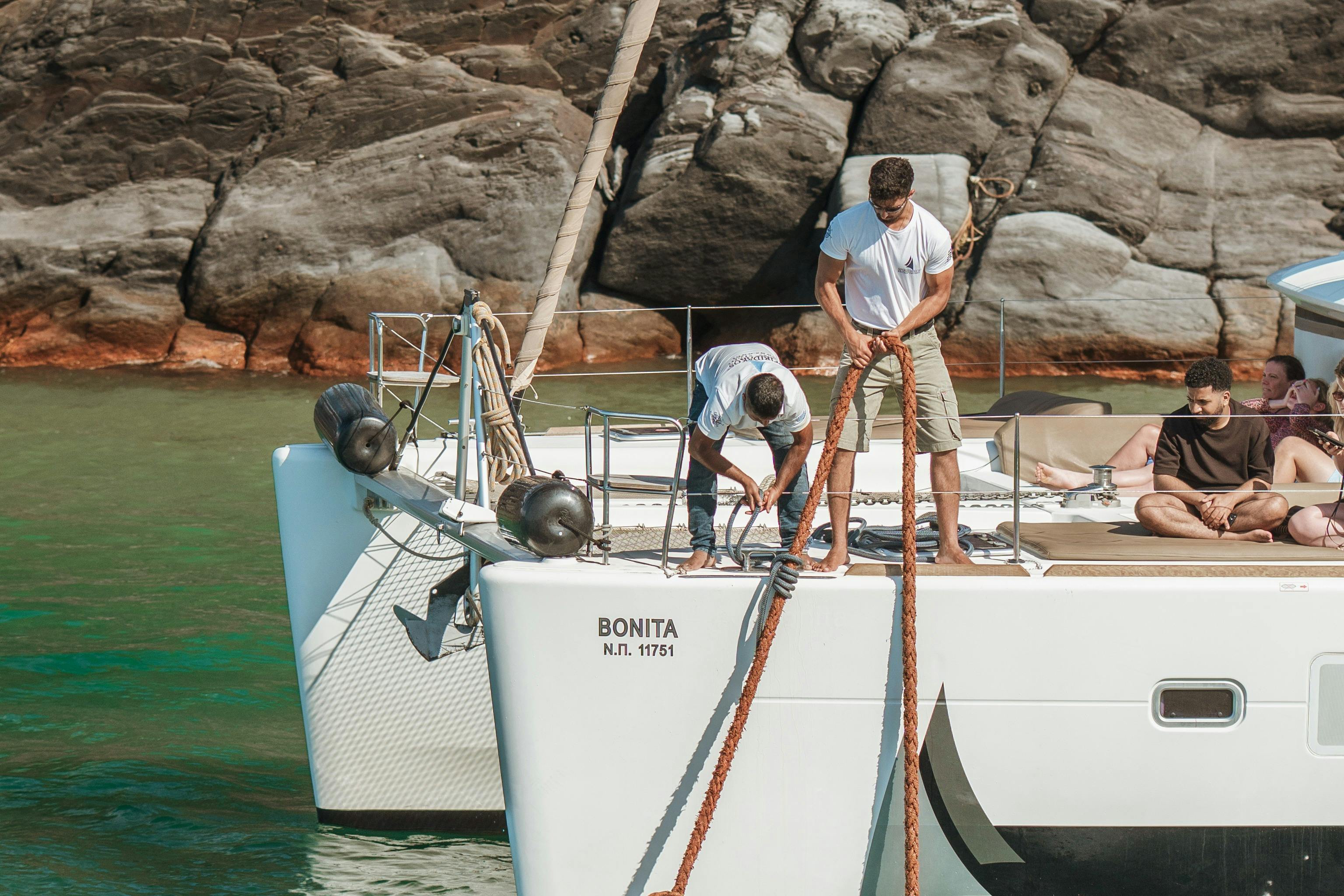 People on a white boat by rocky shores, with two individuals handling ropes and others relaxing on the deck.