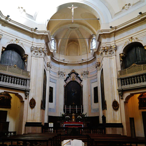 Utsmyckad kyrkinredning med två piporglar, ett altare med målningar, träbänkar och ett stort krucifix som hänger i taket.