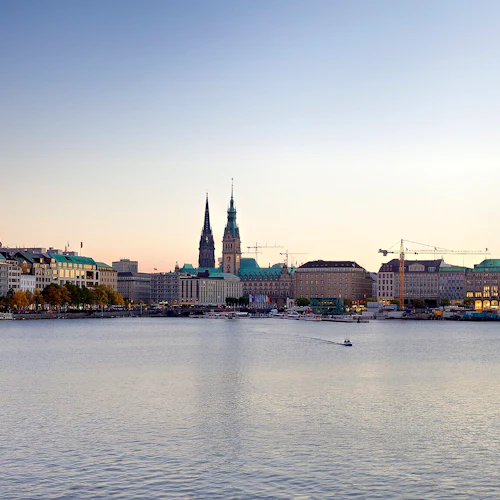 Die Uferpromenade der Stadt in der Abenddämmerung mit historischen Gebäuden mit Türmen, einem Springbrunnen, angedockten Booten und Baukränen.