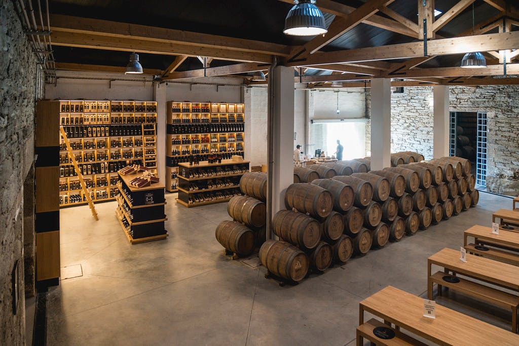 Een opslagruimte van een wijnmakerij met aan de ene kant opgestapelde houten vaten en aan de andere kant planken met uitgestalde wijnflessen.