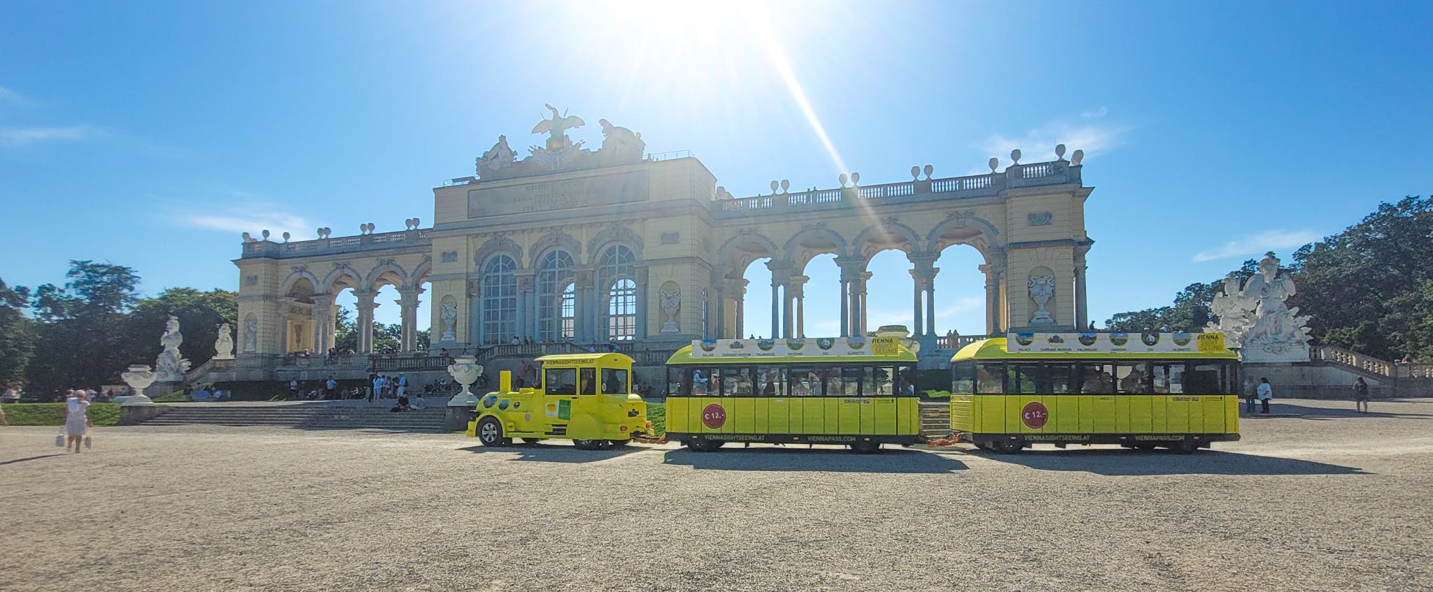 Ett gult turisttåg sitter framför en storslagen, historisk byggnad med valv och statyer under en ljus, solig himmel.