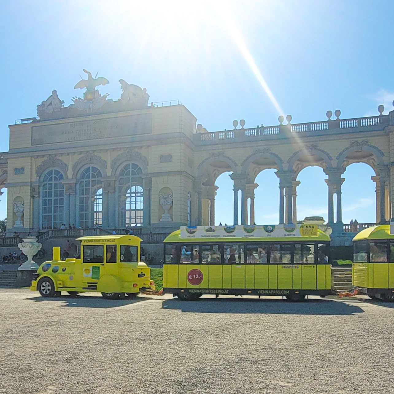 A yellow tourist train sits in front of a grand, historic building with arches and statues under a bright, sunny sky.