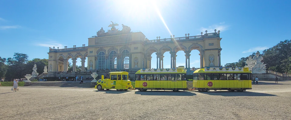 A yellow tourist train sits in front of a grand, historic building with arches and statues under a bright, sunny sky.