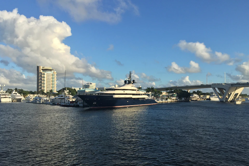 A luxury yacht is docked in a marina with nearby buildings and a bridge under a partly cloudy blue sky.
