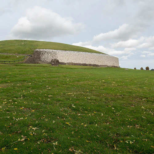 En stor grästäckt kulle med en stenmur, omgiven av öppna gröna fält och en delvis molnig himmel.