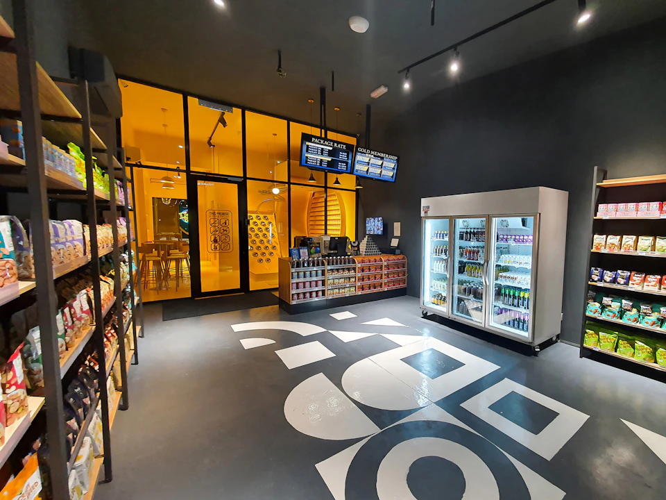 A modern convenience store with shelves of snacks, a refrigerated drinks section, and a bright yellow seating area in the back.