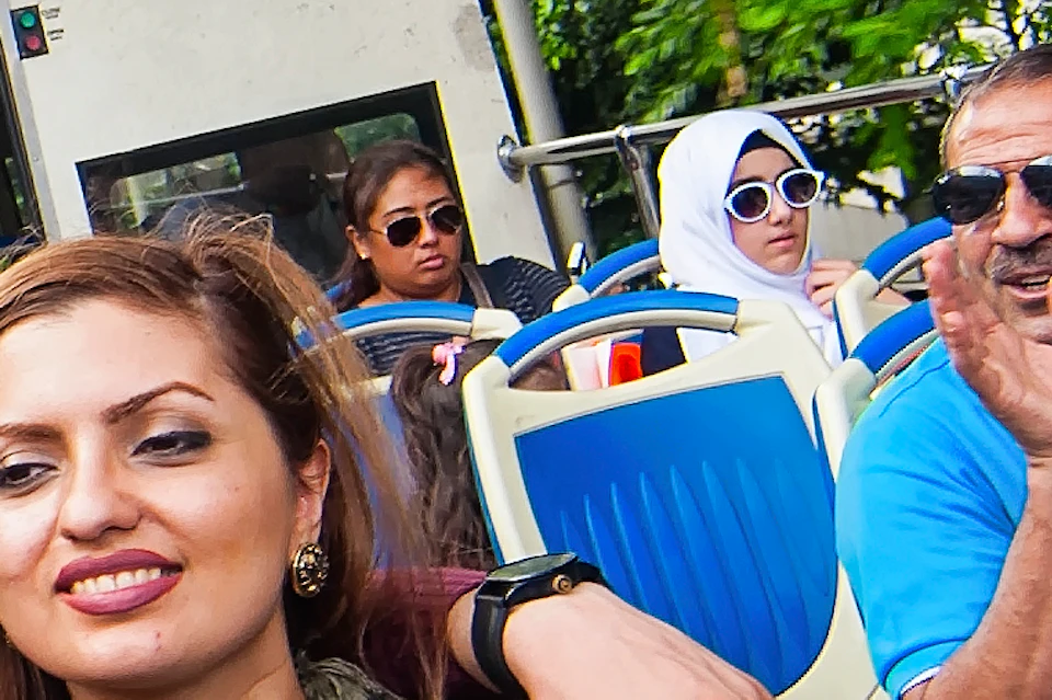 People riding on an open-top tour bus, smiling and surrounded by lush greenery and city structures in the background.