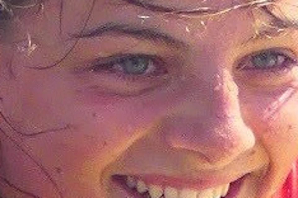 A person in a red wetsuit smiles while lying on a surfboard in shallow water.