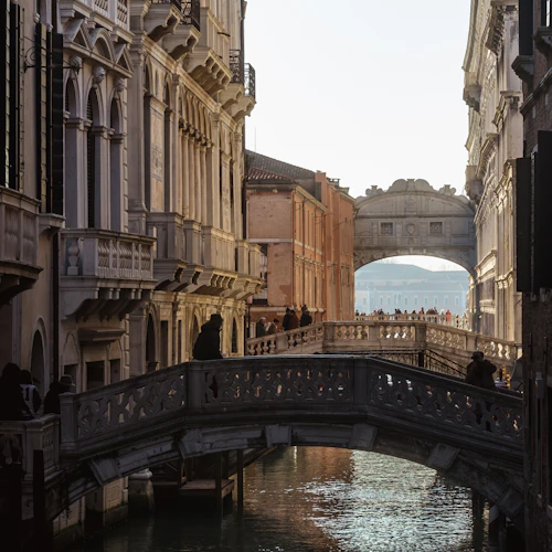 Smal kanal i Venedig med stenbroar och historiska byggnader på båda sidor. Människor går över broarna och längs kanterna.