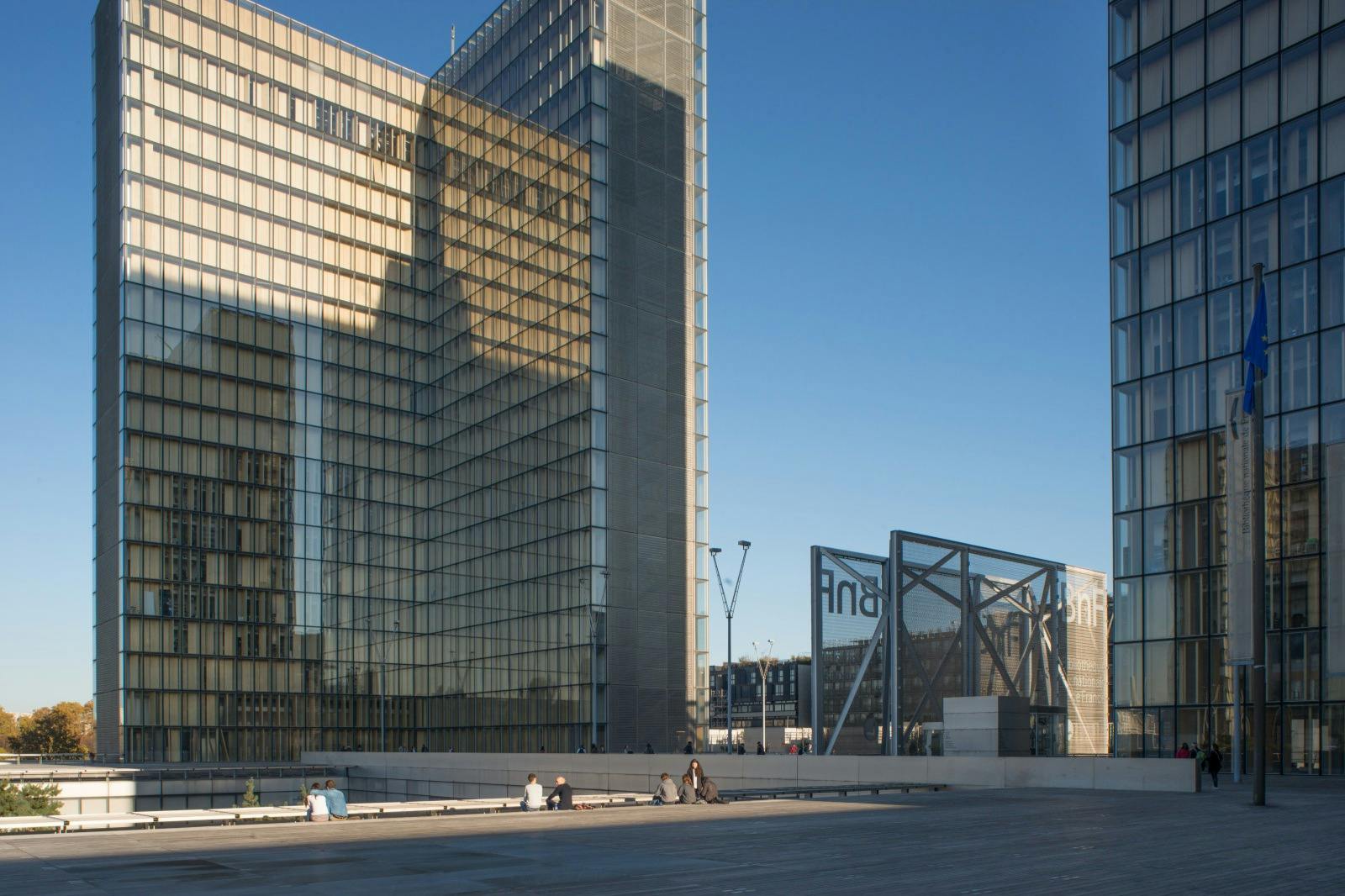Moderna glasbyggnader med människor som sitter på en träplaza under en klarblå himmel.
