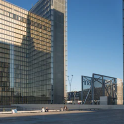 Moderna glasbyggnader med människor som sitter på en träplaza under en klarblå himmel.