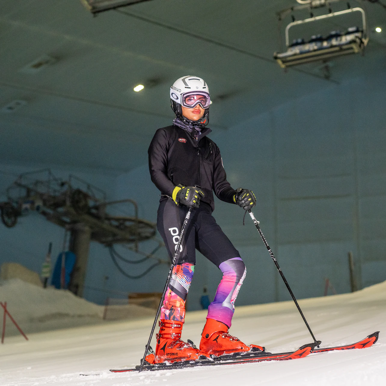 A person wearing ski gear stands on an indoor ski slope, holding ski poles, with a ski lift in the background.