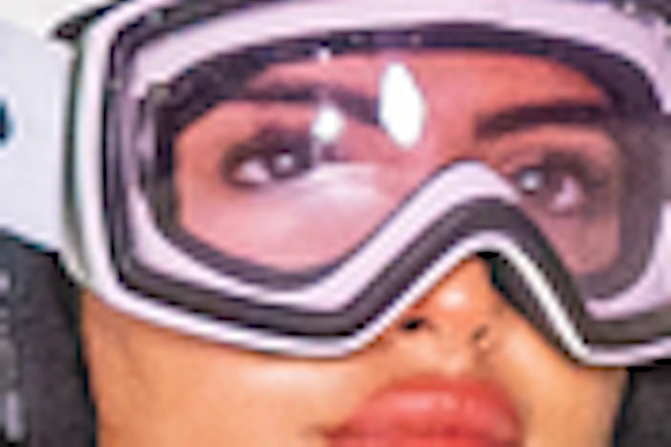 A person wearing ski gear stands on an indoor ski slope, holding ski poles, with a ski lift in the background.