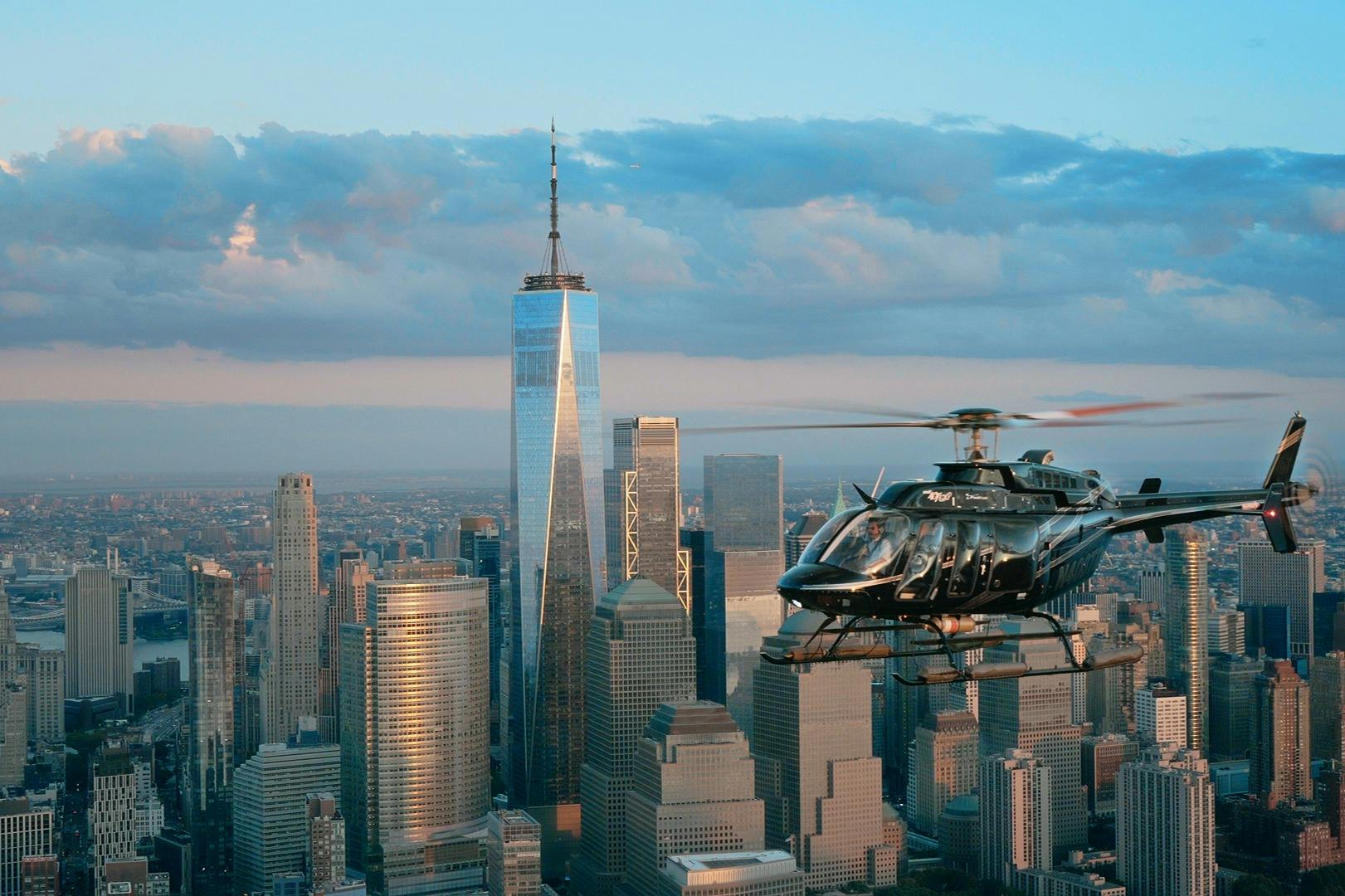 Um helicóptero sobrevoa Manhattan com o One World Trade Center e os arranha-céus circundantes em pano de fundo, sob um céu parcialmente nublado.