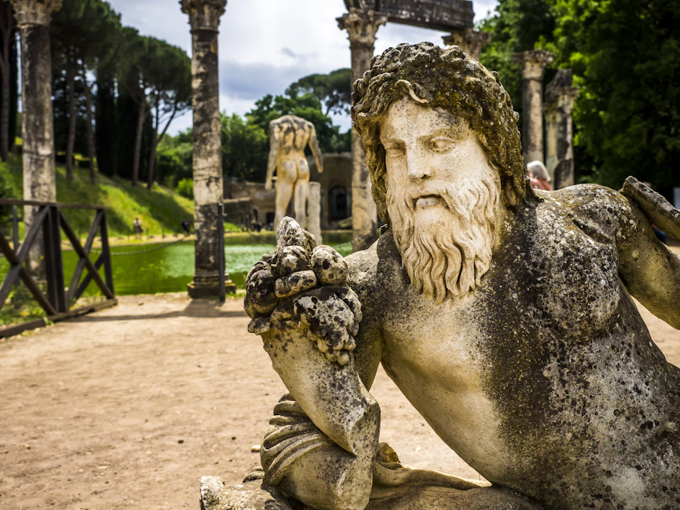 Ancient stone statue of a bearded man in a park, surrounded by columns and greenery with another statue visible in the background.