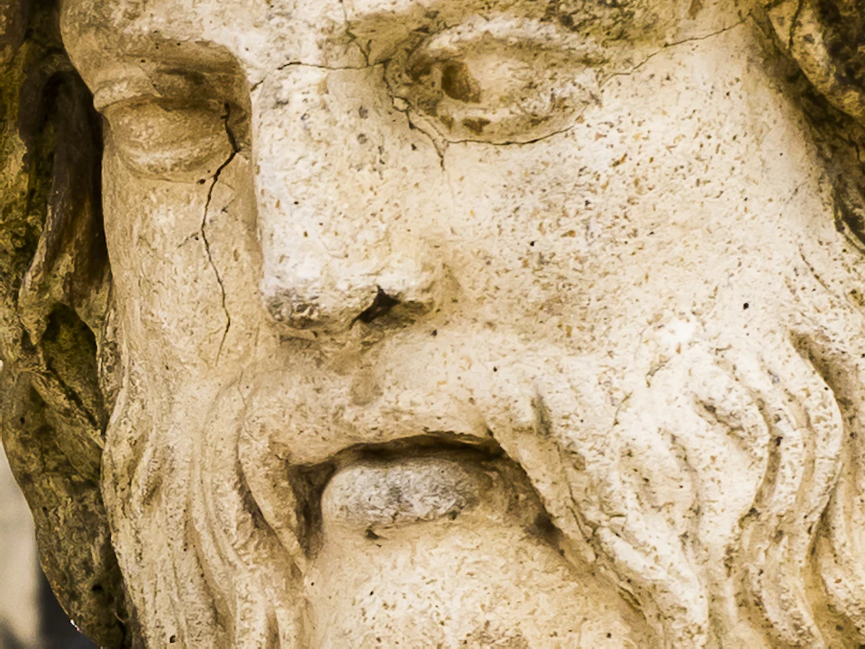 Ancient stone statue of a bearded man in a park, surrounded by columns and greenery with another statue visible in the background.
