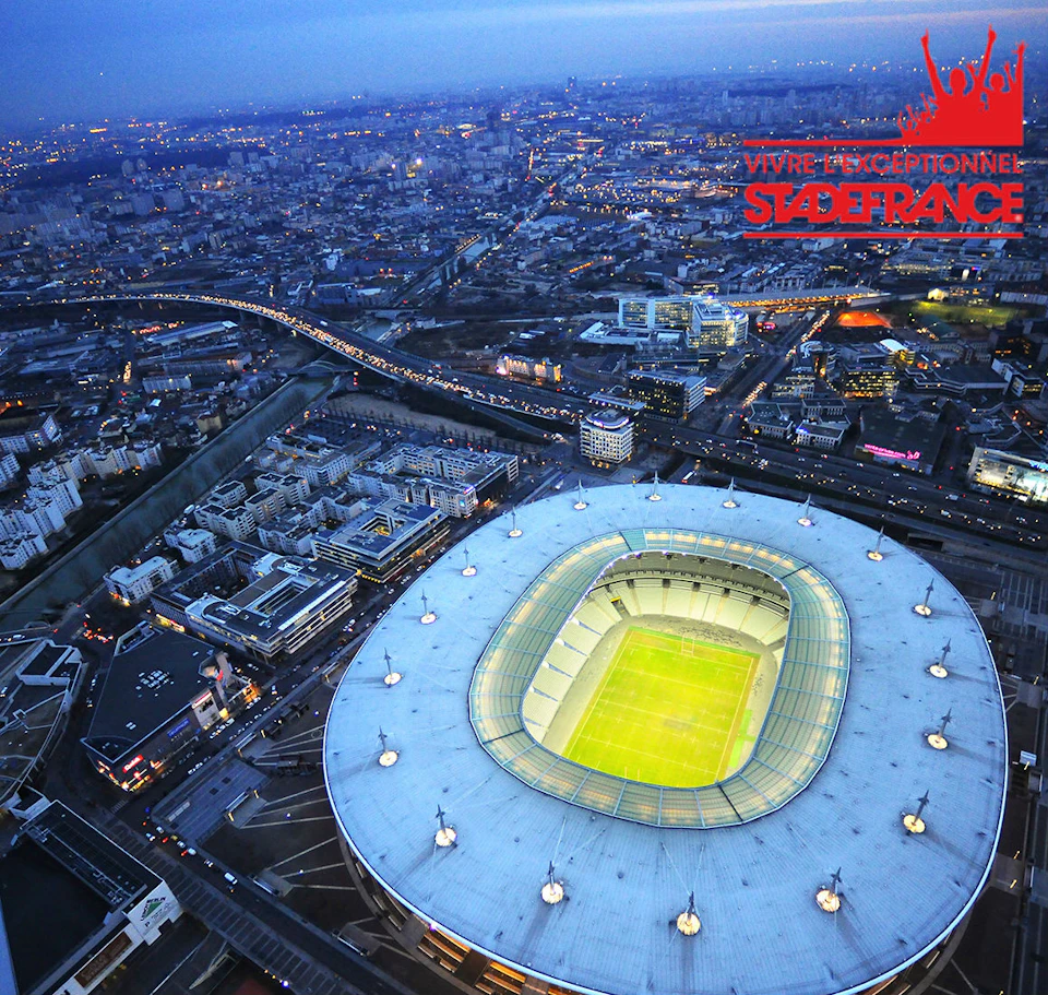 Aerial view of a brightly lit stadium in a bustling city at dusk with roads, buildings, and a river visible.