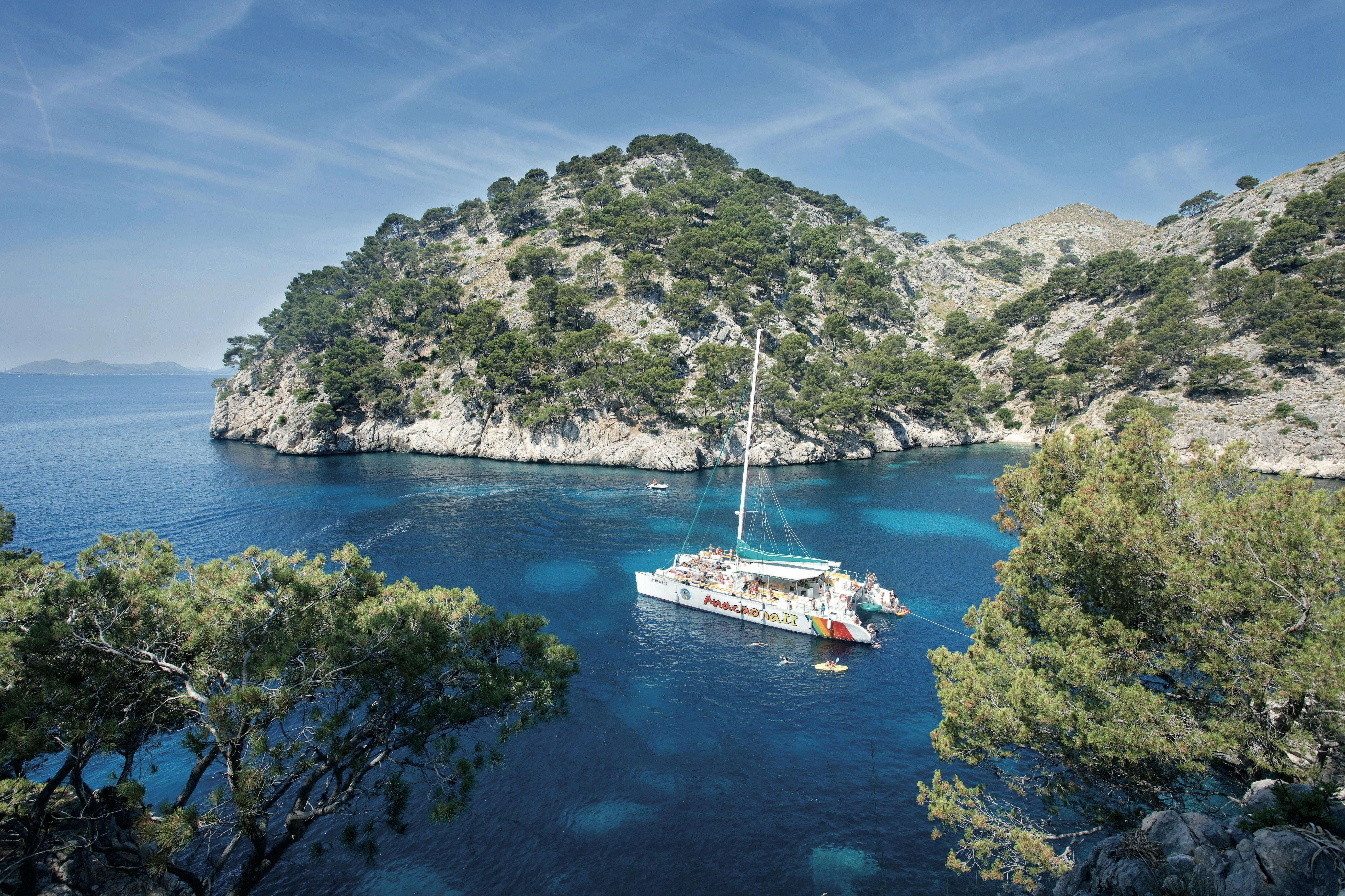 Una barca a vela ancorata in una baia azzurra e limpida, circondata da colline rocciose e ricoperte di alberi, sotto un cielo limpido.