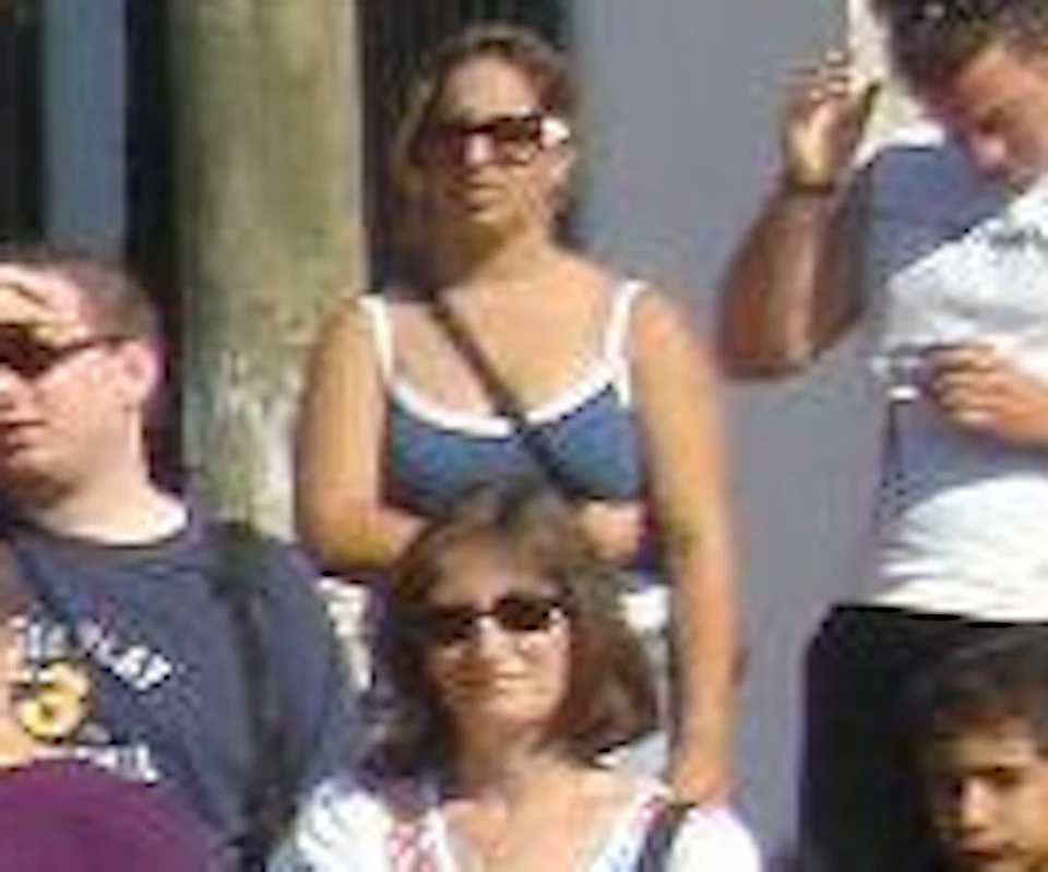 A brown camel stands on a dirt path, surrounded by a small crowd of people near white buildings under a clear blue sky.