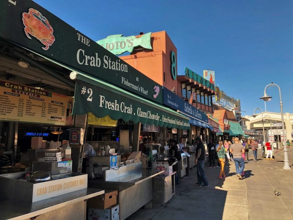People walking past a row of outdoor seafood vendors at Fisherman's Wharf, including "The Crab Station." Signs advertise fresh crab and clam chowder.