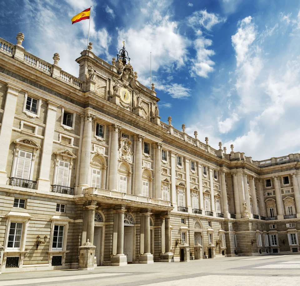 Ornate palace with Spanish flag on rooftop, numerous windows, columns, and detailed facade under a partly cloudy blue sky.