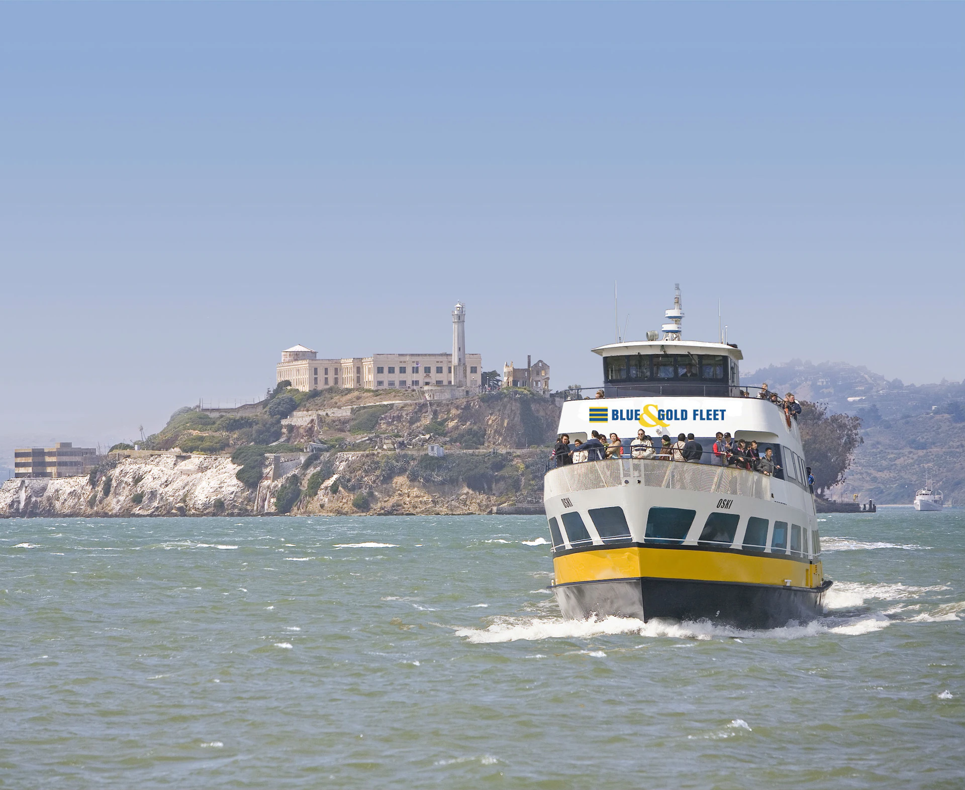 A ferry labeled "Blue & Gold Fleet" approaches in the foreground with an island and historic-looking buildings in the background.