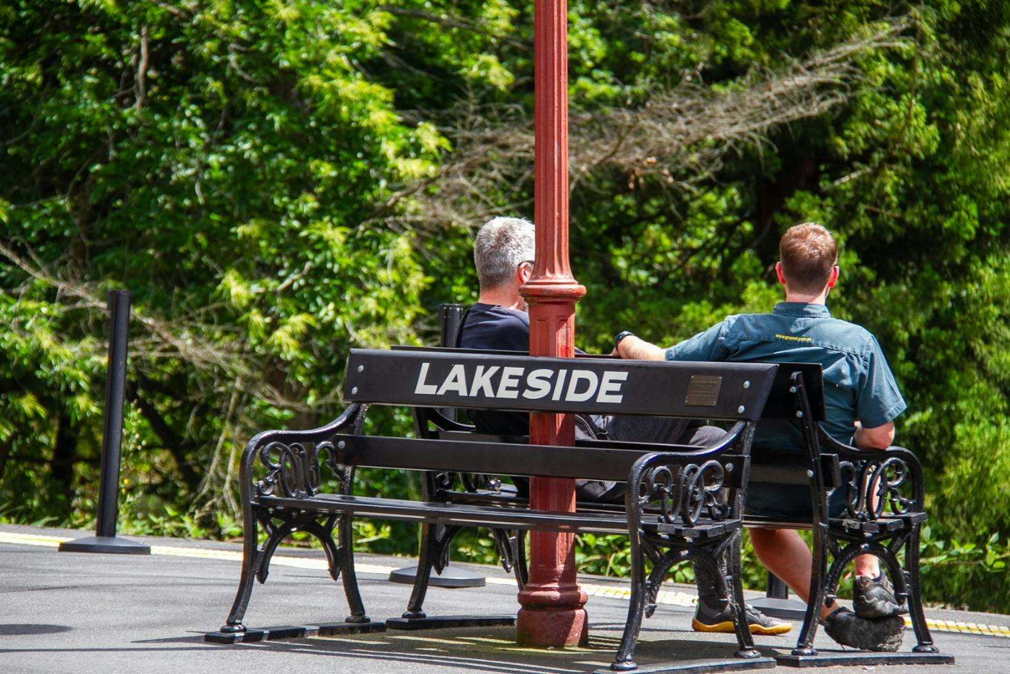 Two people sitting on a bench labeled "Lakeside" near a red pole, with green trees in the background.