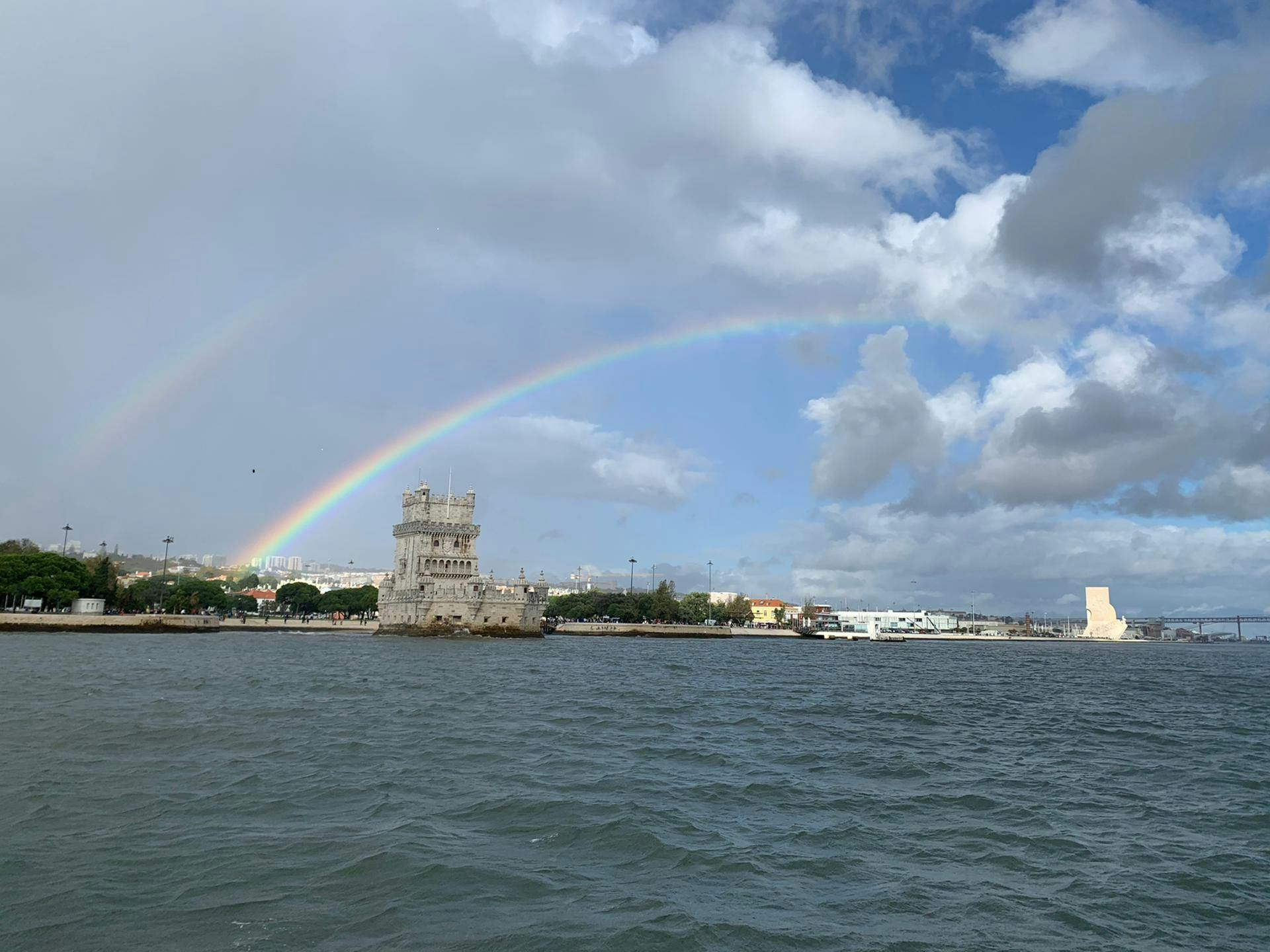Una storica torre di pietra si erge sul lungomare sotto un cielo parzialmente nuvoloso, con un doppio arcobaleno che la sovrasta.
