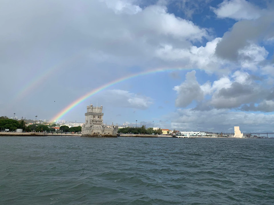 A historic stone tower stands by the waterfront under a partly cloudy sky, with a double rainbow arching above it.