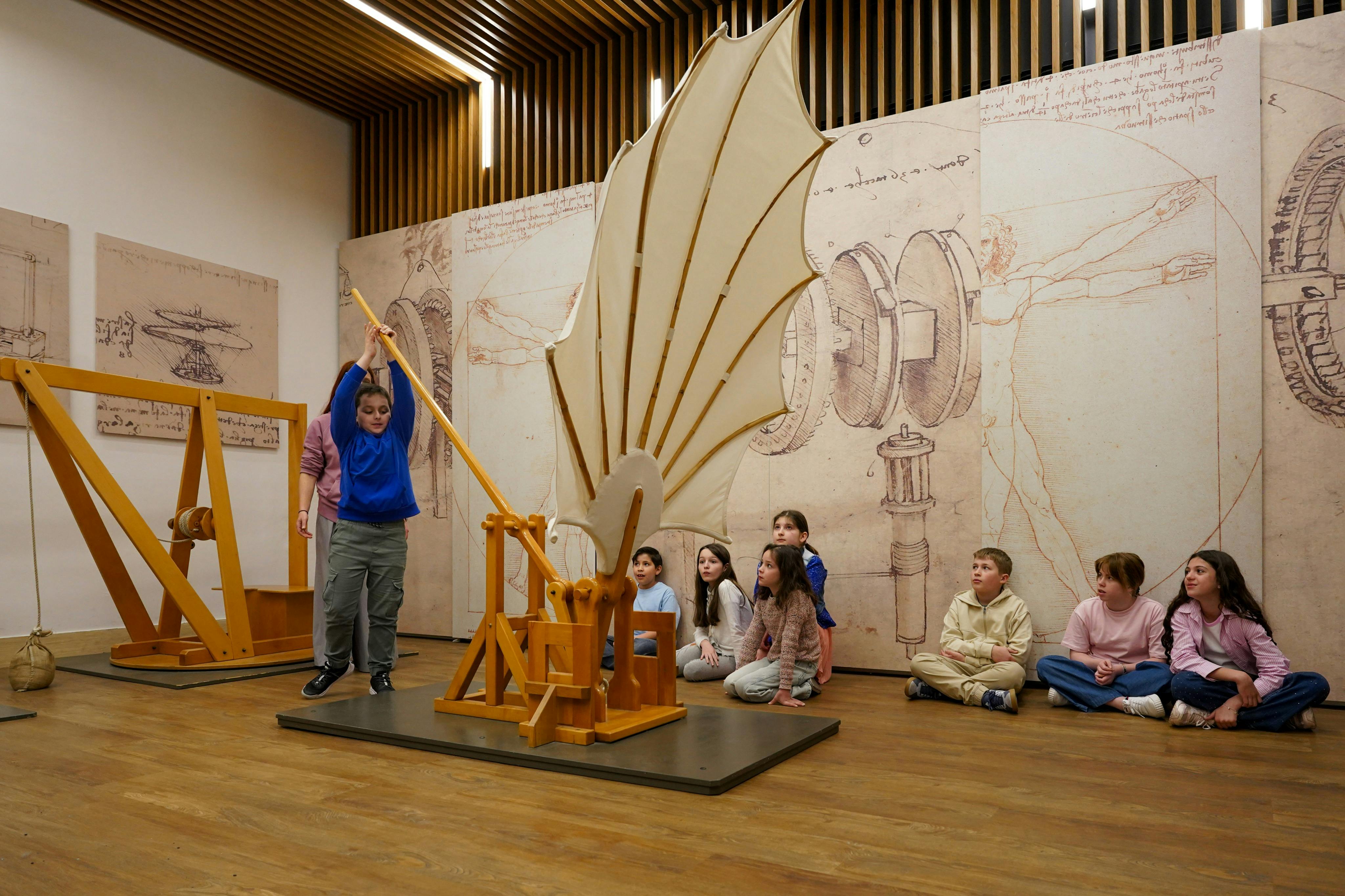 I bambini interagiscono con i modelli in legno delle invenzioni di Leonardo da Vinci in una mostra museale con schizzi appesi alle pareti.
