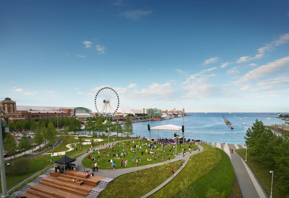 People enjoying a park with a Ferris wheel, grass lawn, stage, and waterfront view under a clear blue sky.