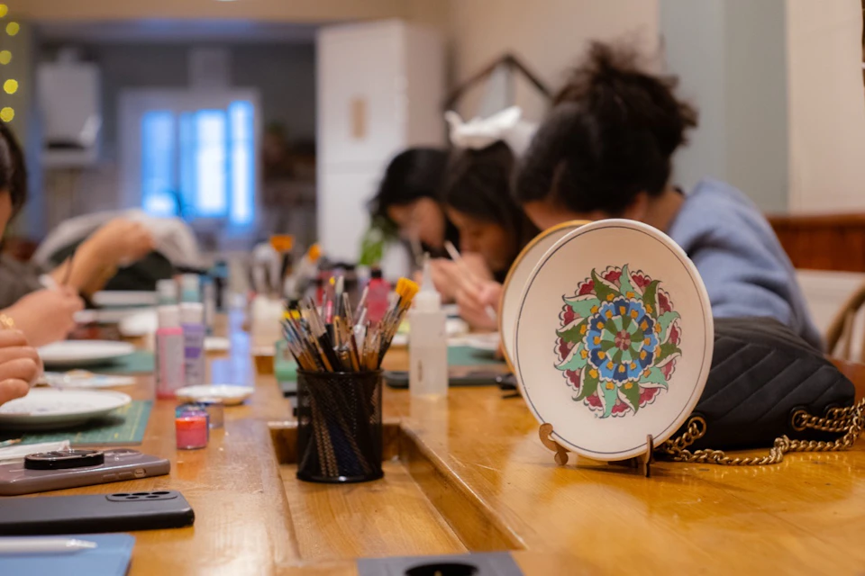 People sitting at a table engaged in painting crafts, with brushes and art supplies scattered. A decorated plate is displayed.