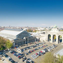 Flygfoto över en stor byggnad med glastak och intilliggande båge, omgiven av parkerade bilar och träd, belägen i en stadsbild.