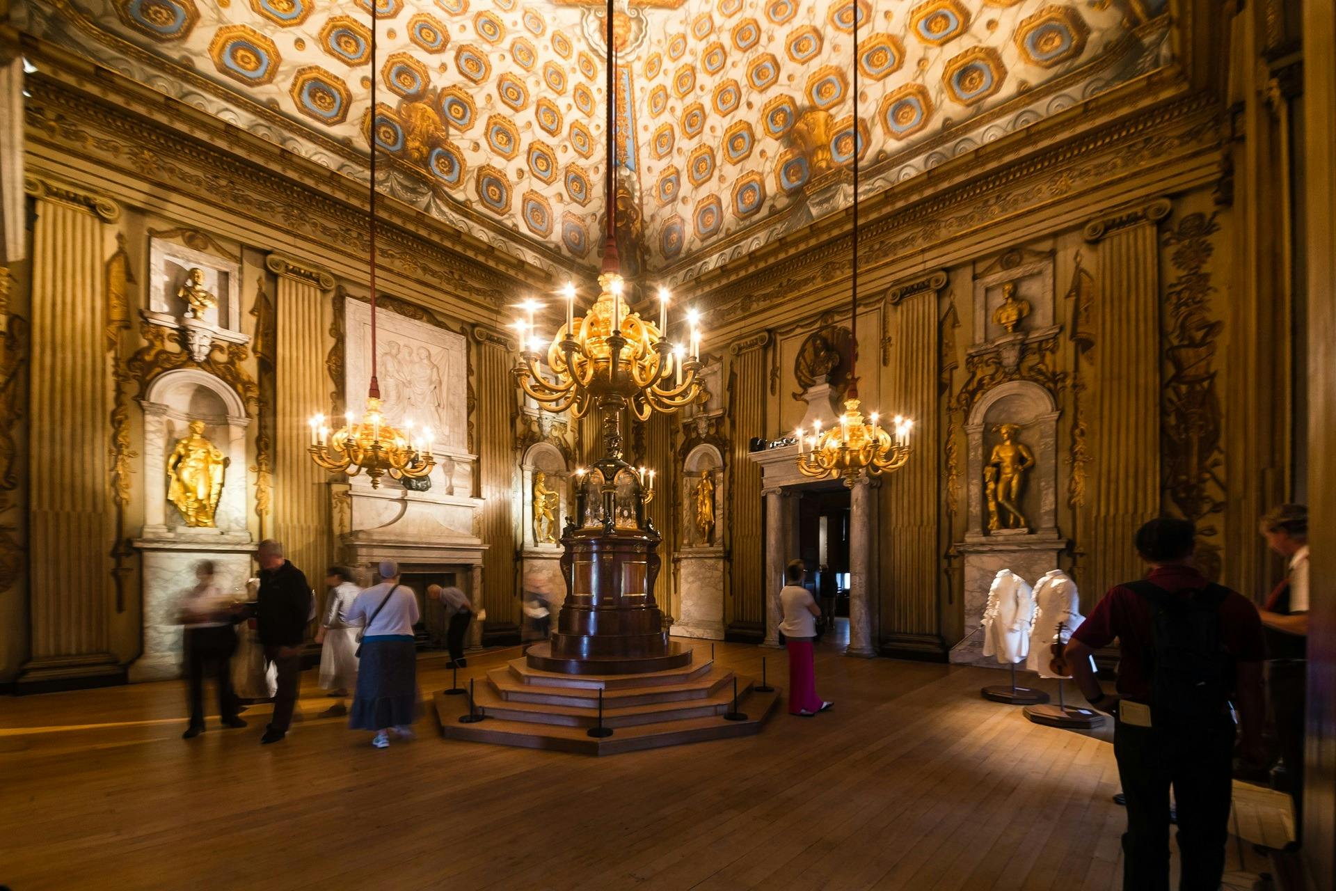 Ornate room with decorated ceiling, chandeliers, gold statues, and visitors observing artworks and displays.