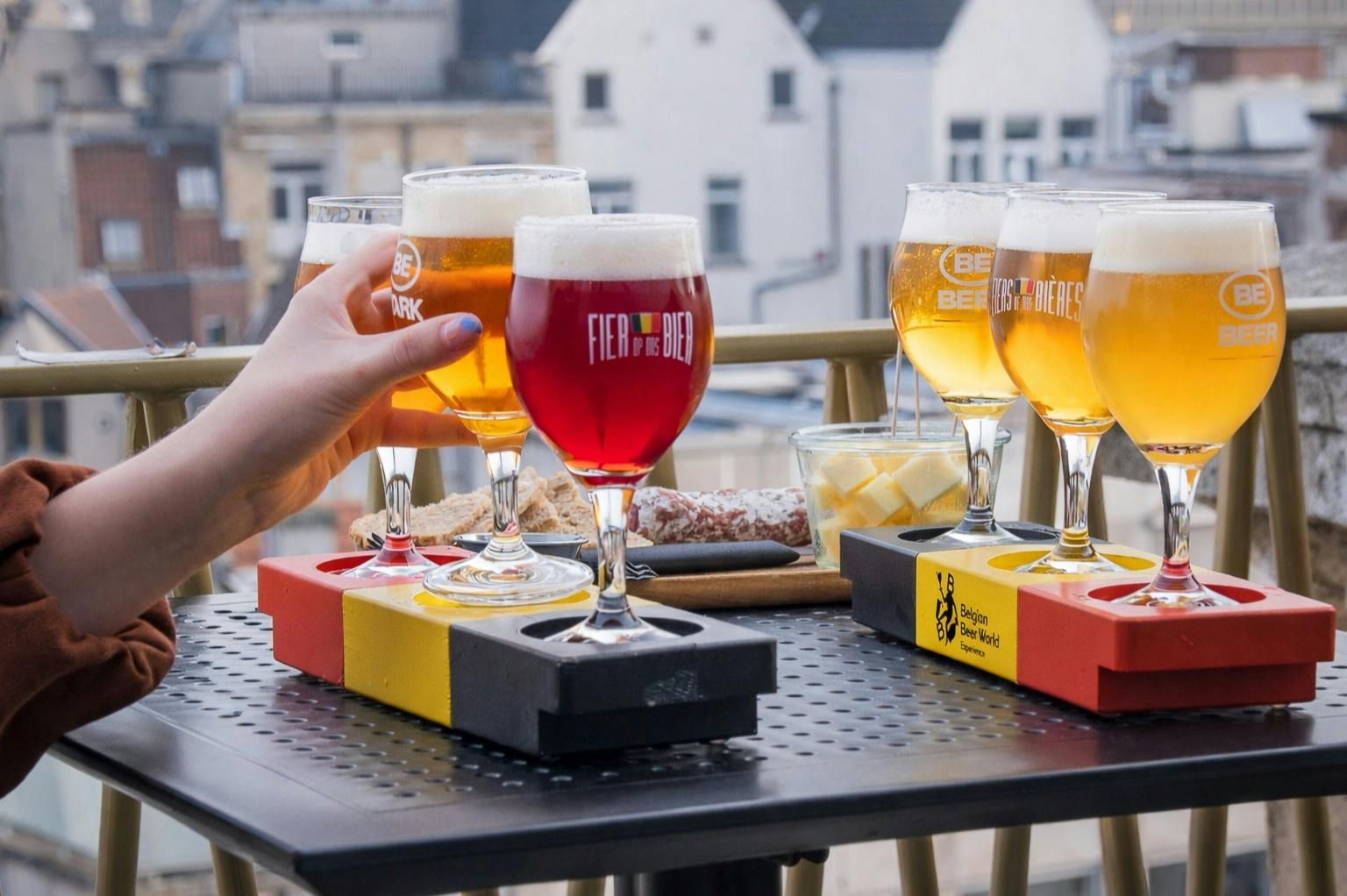 A table with glasses of beer in flight holders, a hand raising one glass, and food items in the background on a terrace.