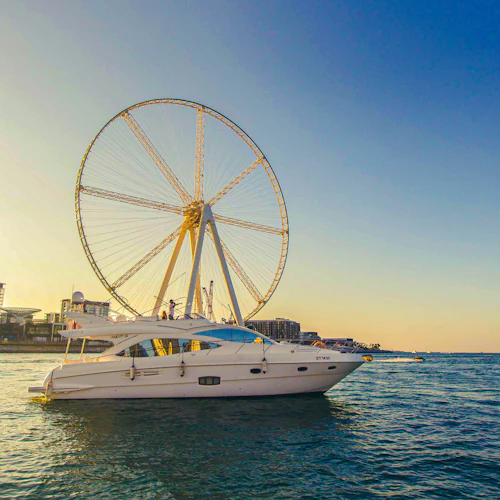 Eine weiße Jacht auf dem Wasser mit einem riesigen Riesenrad und modernen Gebäuden im Hintergrund während der goldenen Stunde.