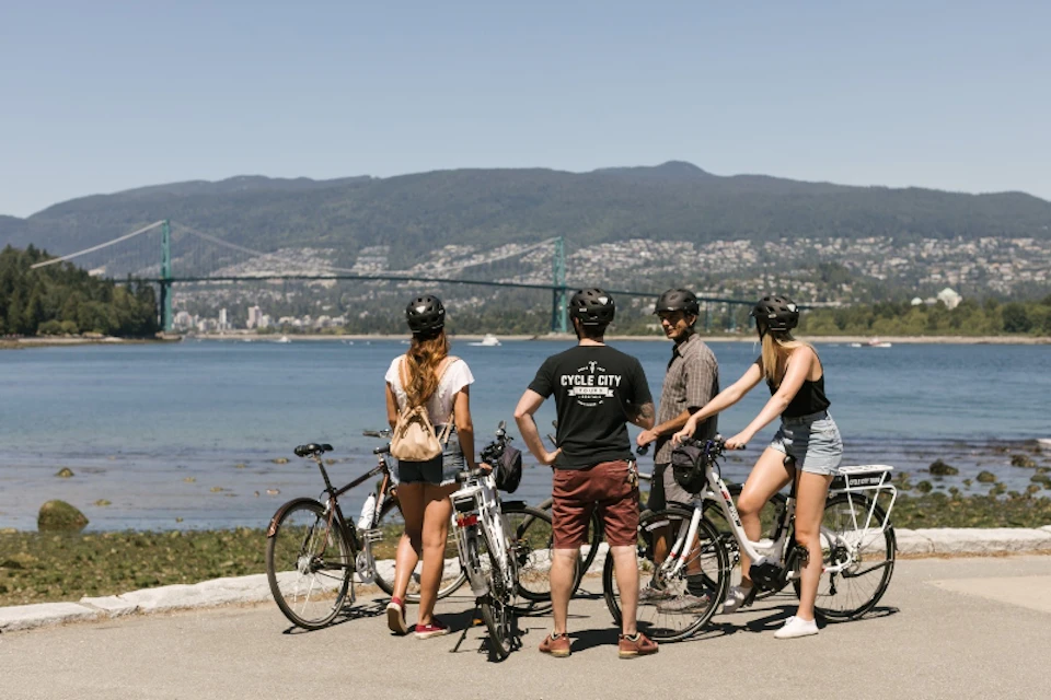 Four people with bicycles stand by a waterfront, looking towards a cityscape with a bridge and mountains in the background.
