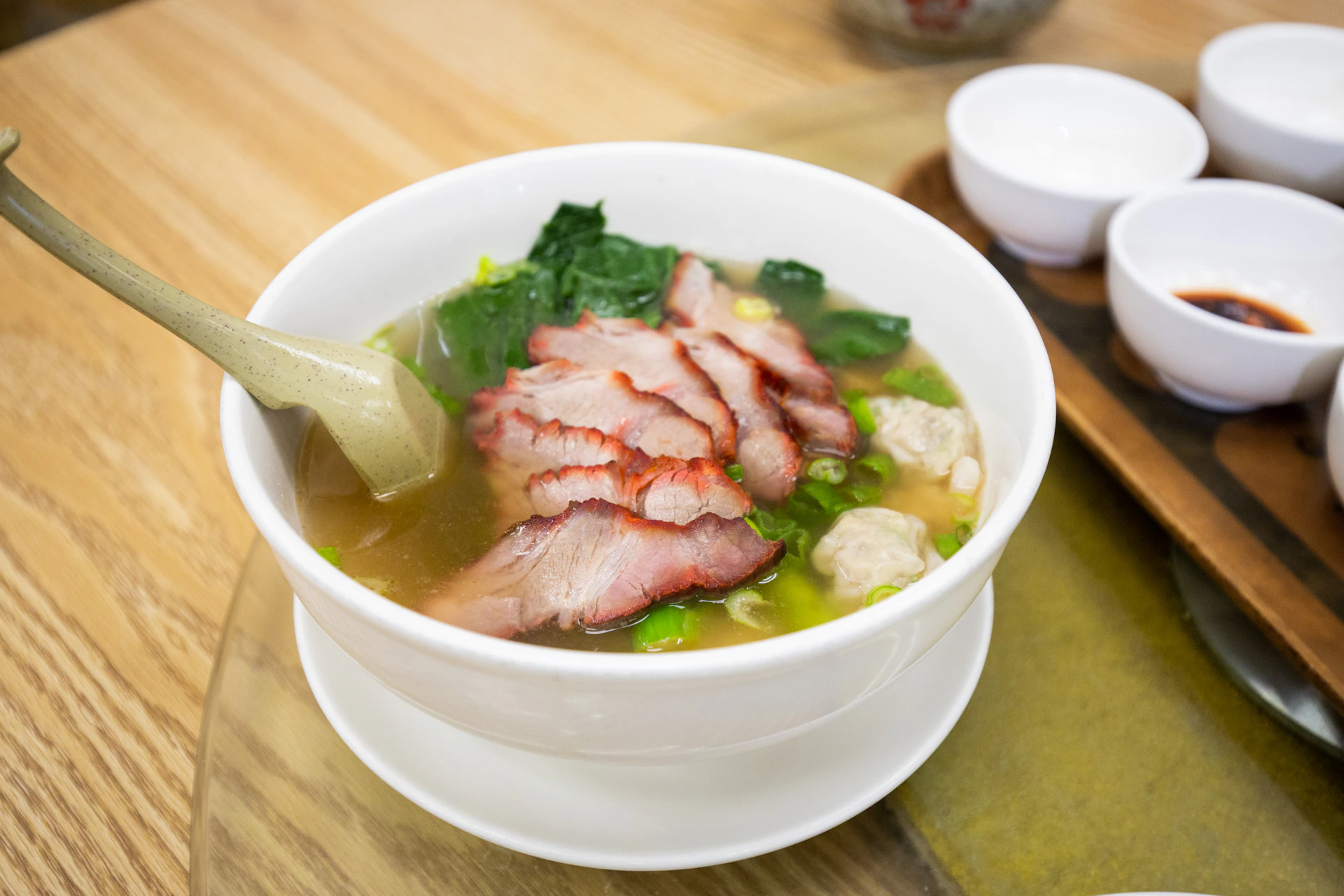 A bowl of noodle soup with sliced pork, green vegetables, and meatballs, served with a ceramic soup spoon.