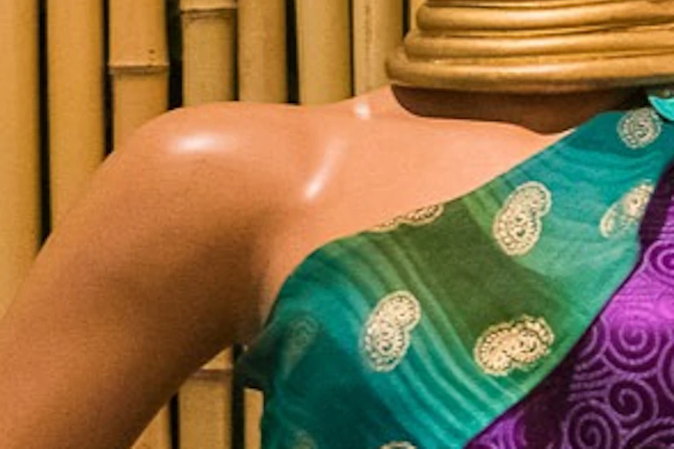 A mannequin dressed in traditional attire with neck rings sits on a wooden bench against a bamboo backdrop.